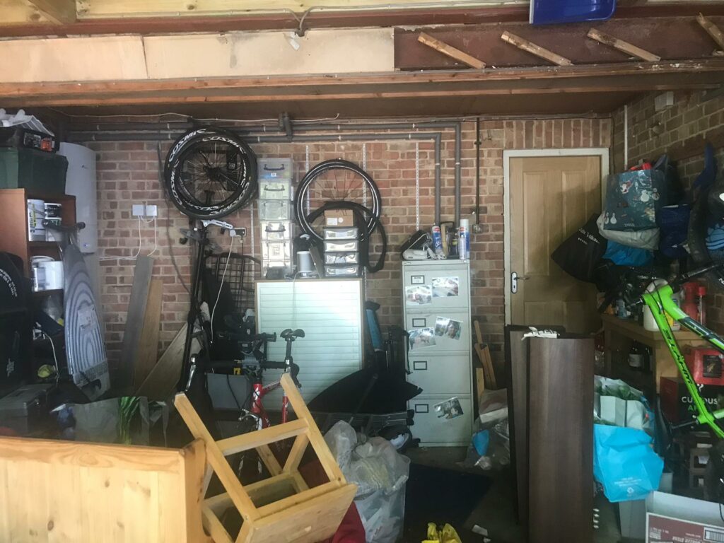A messy & dark garage, cluttered with tables, chairs and bikes.