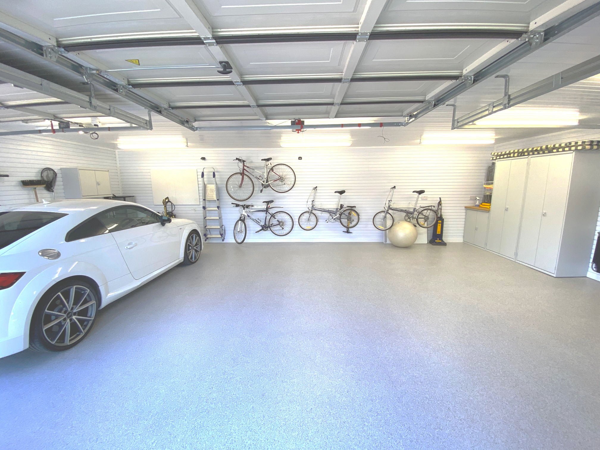 A double garage with a blue resin floor, white cladded walls, bike racks and a white car on the left of the image.