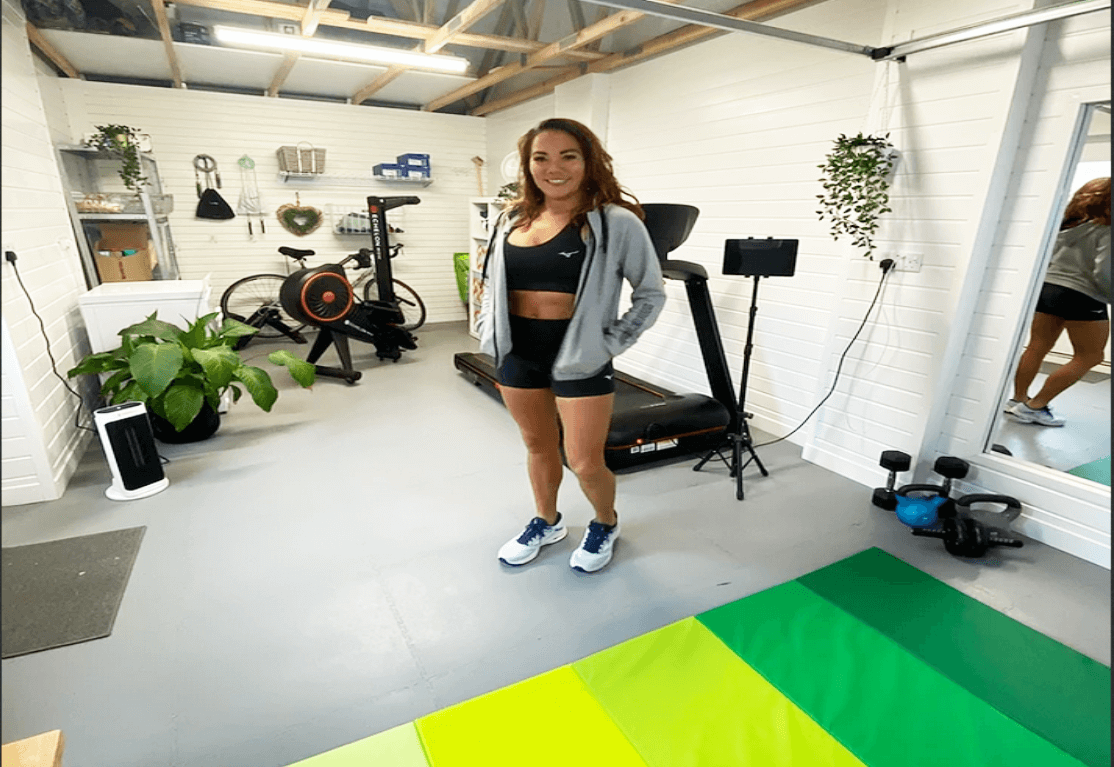 A garage converted into a gym with a woman standing in the middle of the room and a treadmill in the background.