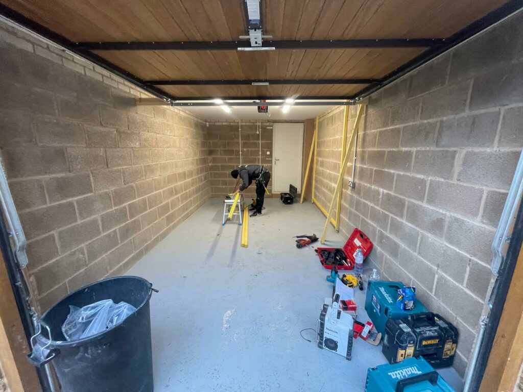 The inside of a garage with a man stood in the middle, measuring wooden planks.